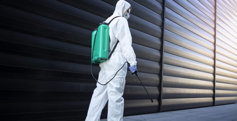 Shot of exterminator person in white chemical protection suit doing disinfection and pest control with sprayer to kill insects and rodents.